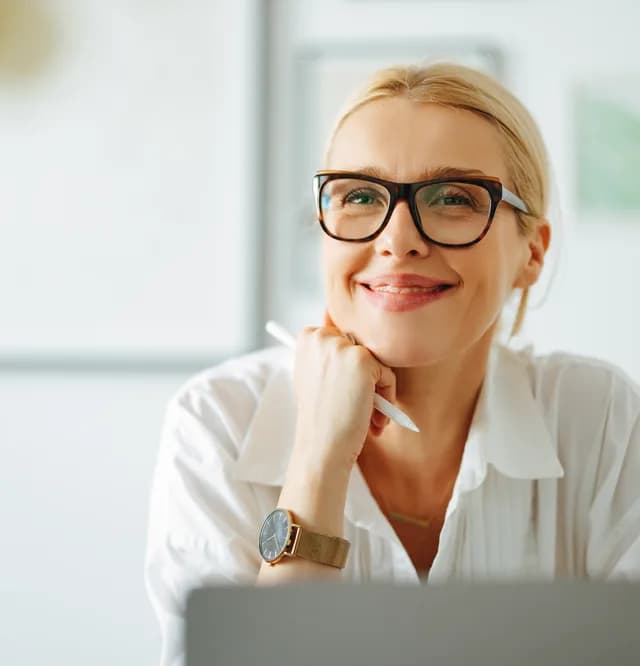 Frau mit Brille sitzt lächelnd mit Stift vor einem Laptop