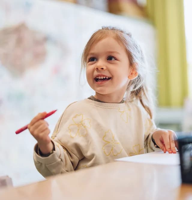 Kleines Mädchen malt lachend mit einem roten Stift am Tisch